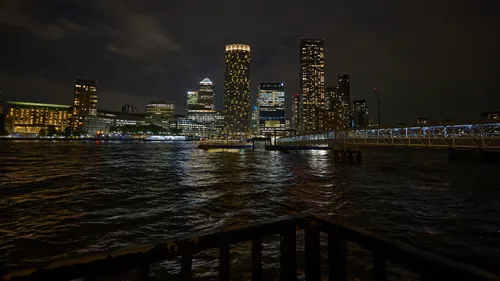 Canary Wharf night pier This urban night photograph shows Canary Wharf in London, United Kingdom, as seen from the pier by the River Thames. The scene was captured in the evening during early autumn, showcasing illuminated skyscrapers such as One Canada Square and other prominent buildings that define the Canary Wharf skyline. Reflections from the office towers and riverside lights shimmer across the water, highlighting the vibrant atmosphere along the Thames. The architecture of the area is clearly visible in the image, with the Canary Wharf pier and modern footbridge stretching out toward the high-rise district, illustrating the dynamic urban environment synonymous with central London.